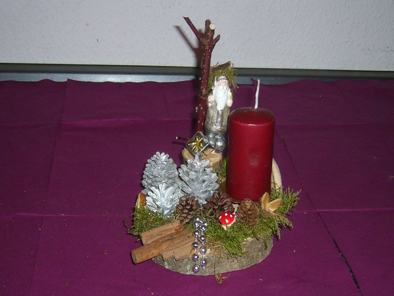 Rundes 1er Gesteck auf Baumscheibe mit dunkelroter Kerze und silbernem Weihnachtsmann, knapp 20 cm im Durchmesser zu 8,00 €.