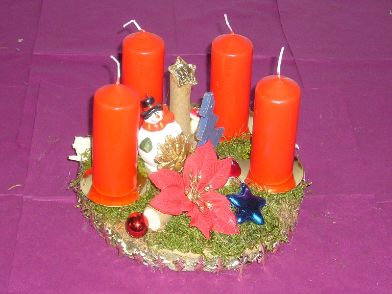 Kleineres 4er Gesteck mit roten Kerzen auf Baumscheibe mit Moos und reichlich Weihnachtsdekoration für 10,00 €. 